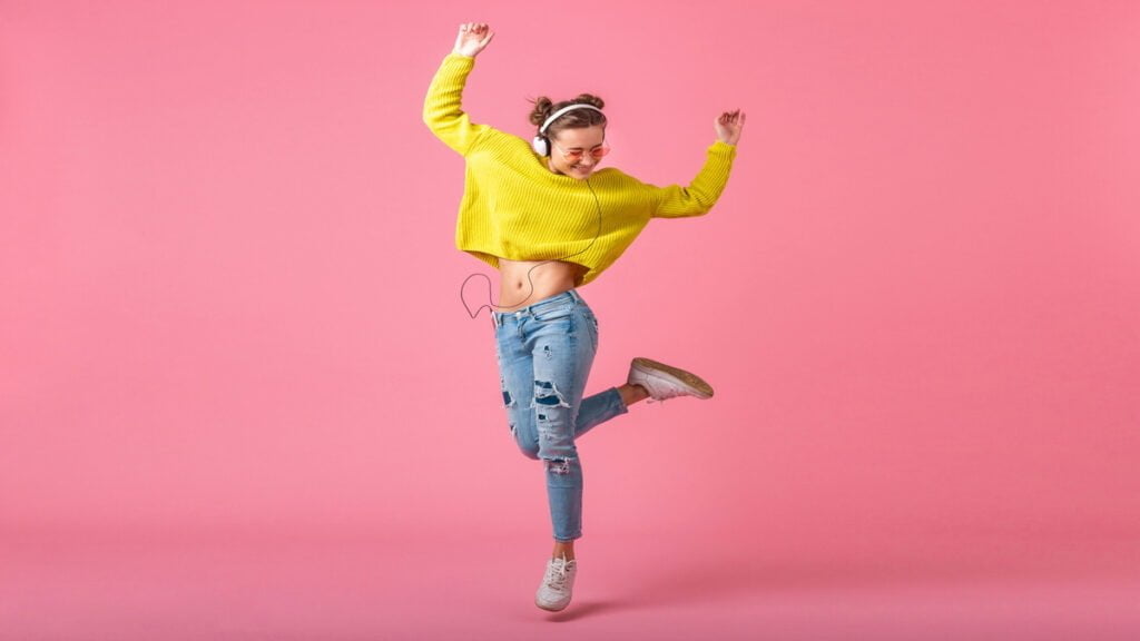 attractive happy funny woman jumping listening to music in headphones dressed in hipster colorful style outfit isolated on pink wall wearing yellow sweater and sunglasses having fun