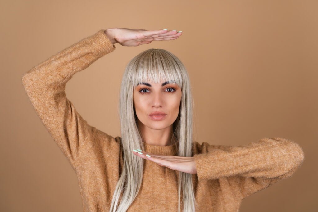 a blonde girl with bangs and brown daytime make up in a sweater on a beige background pulls her hands above her head and under her chin