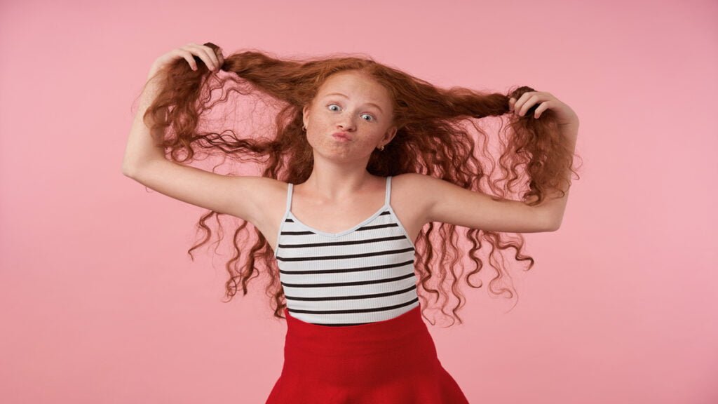 studio shot funny redhead female kid with long curly hair making faces pink background looking camera with raised eyebrows pursing lips wearing festive clothes